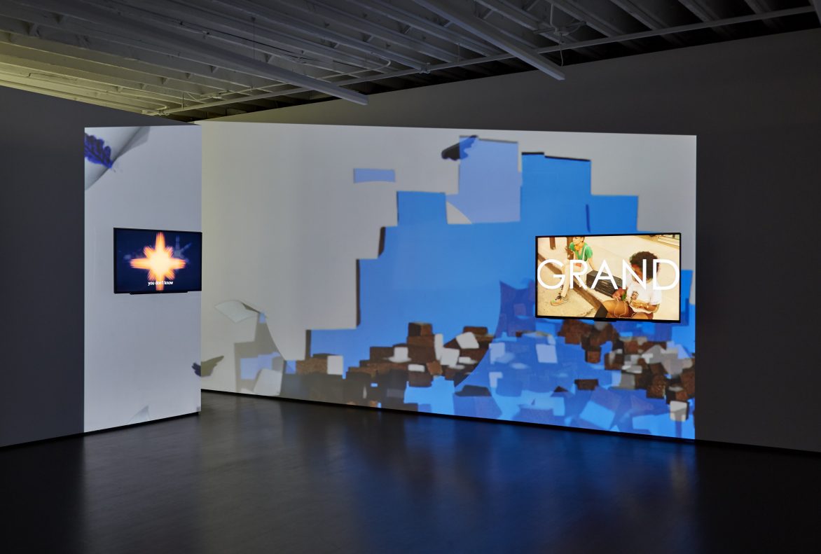 View of a room with projection on two walls of blue square shaped abstraction with a screen mounted in the wall that says 