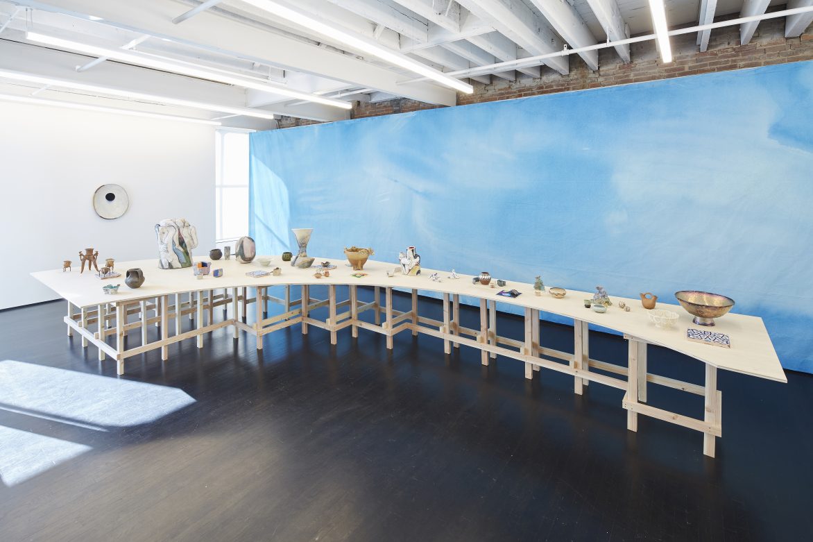 View of a long curved table with ceramic vessels on top, against a wall painted sky blue.
