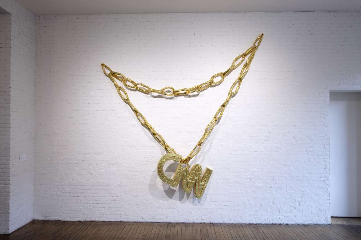 Installation view of an oversized gold chainlink necklace with the letters CNN hanging from chain.