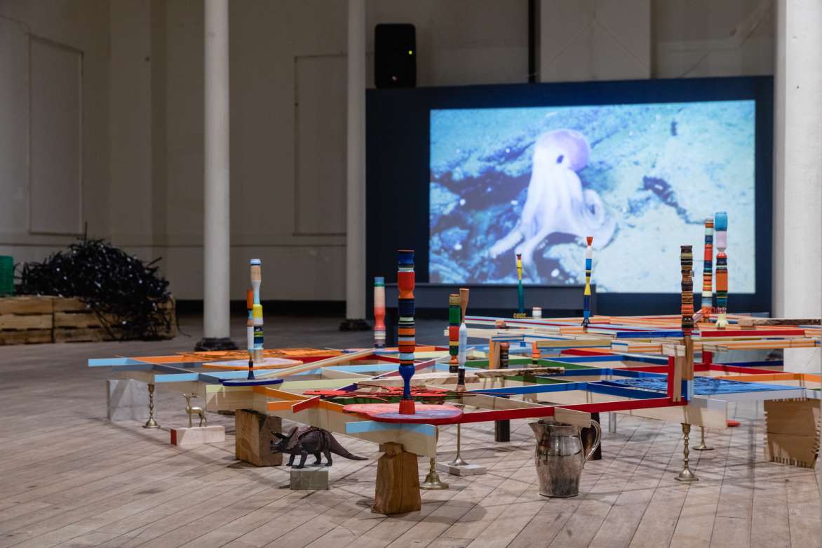 Installation view of wooden grid-like sculpture that is support by various objects with a video projection in the background.
