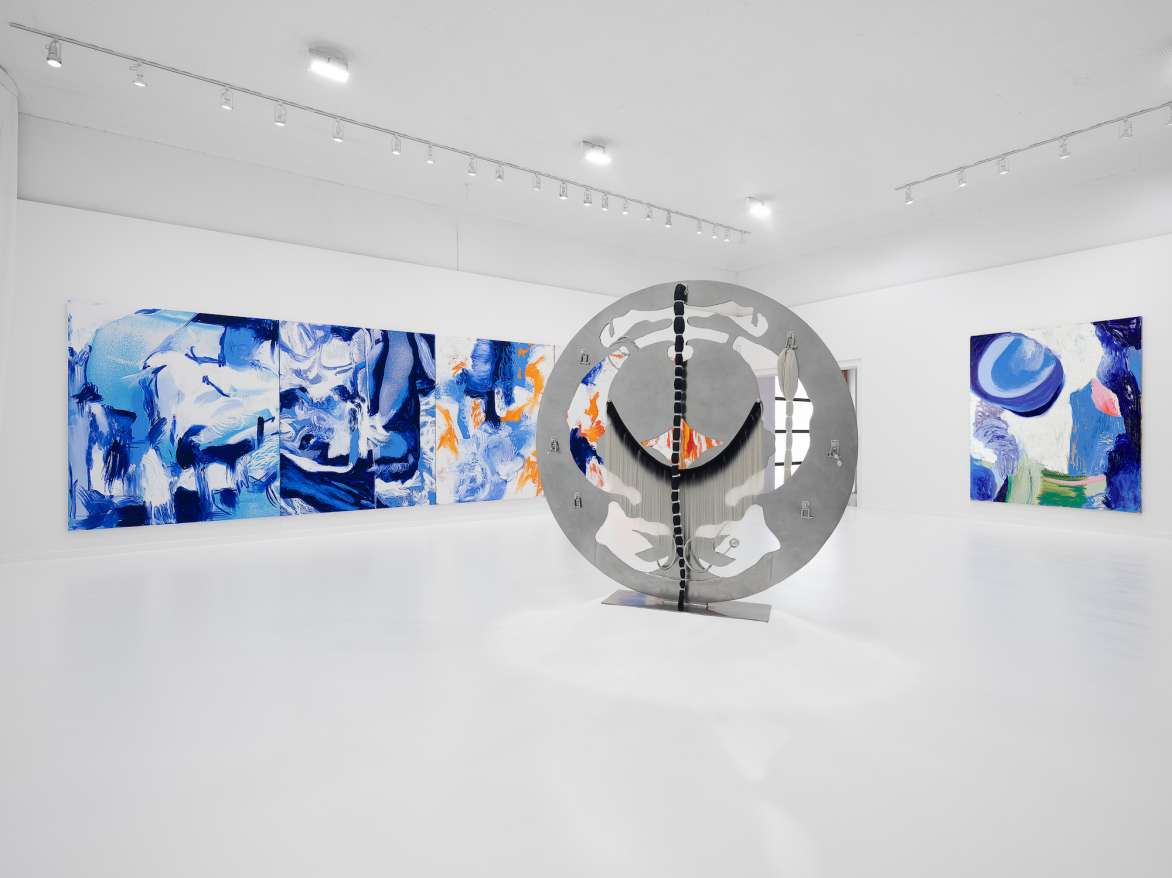 Installation view of a large round steel sculpture in the center of the room with blue and white abstract paintings on the wall.