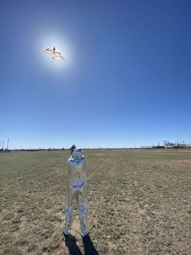 A figure in a silver suit flying a kite in the shape of a bird. The kite is directly in front of the sun.
