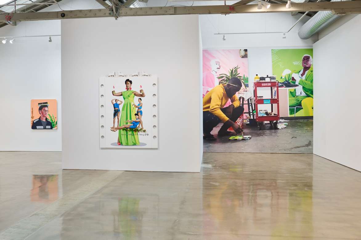 View of a gallery with a larger portrait of a woman in a green dress. To left beyond the wall with the portrait is a mural-size photograph of a man kneeling on the floor dipping a paintbrush into a paint tray.