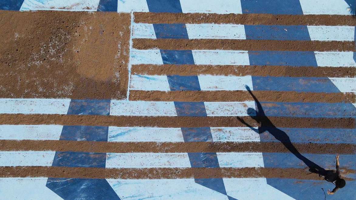 Picture of a concrete ground that has been painting with geomteric shapes in blue and light blue, with paint removed to expose the concrete in the design of the American flag. The shadow of a small figure with arms raised is in the lower left corner.
