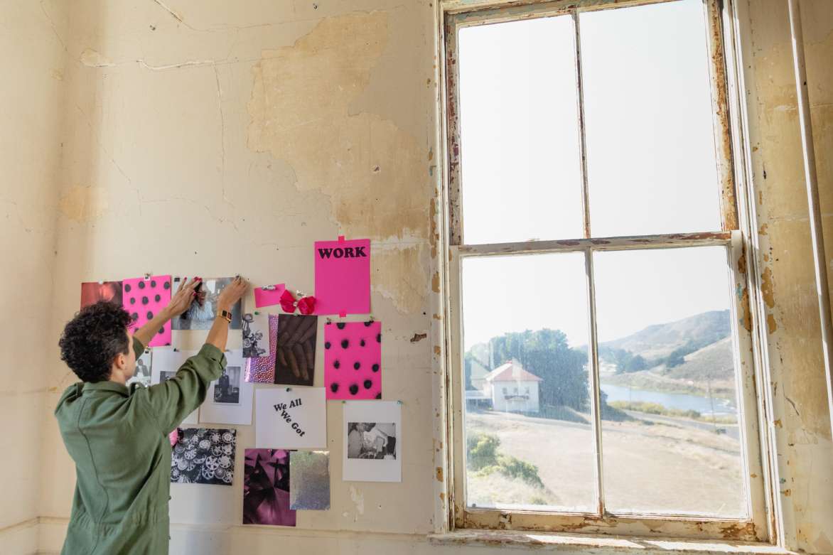 A woman pinning a photograph to the wall where pieces of bright pink paper have been adhered. There is a window to the left with a view of white building and rolling hills behind it.