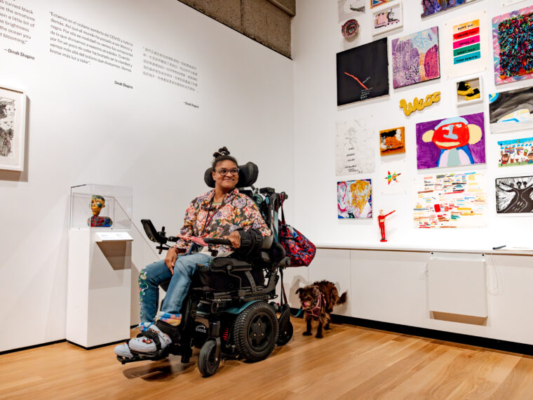 NIAD artist MiaMya Dawson poses in her wheelchair in front of a wall displaying NIAD artwork at Oakland Museum of California’s “Into the Brightness” exhibition.