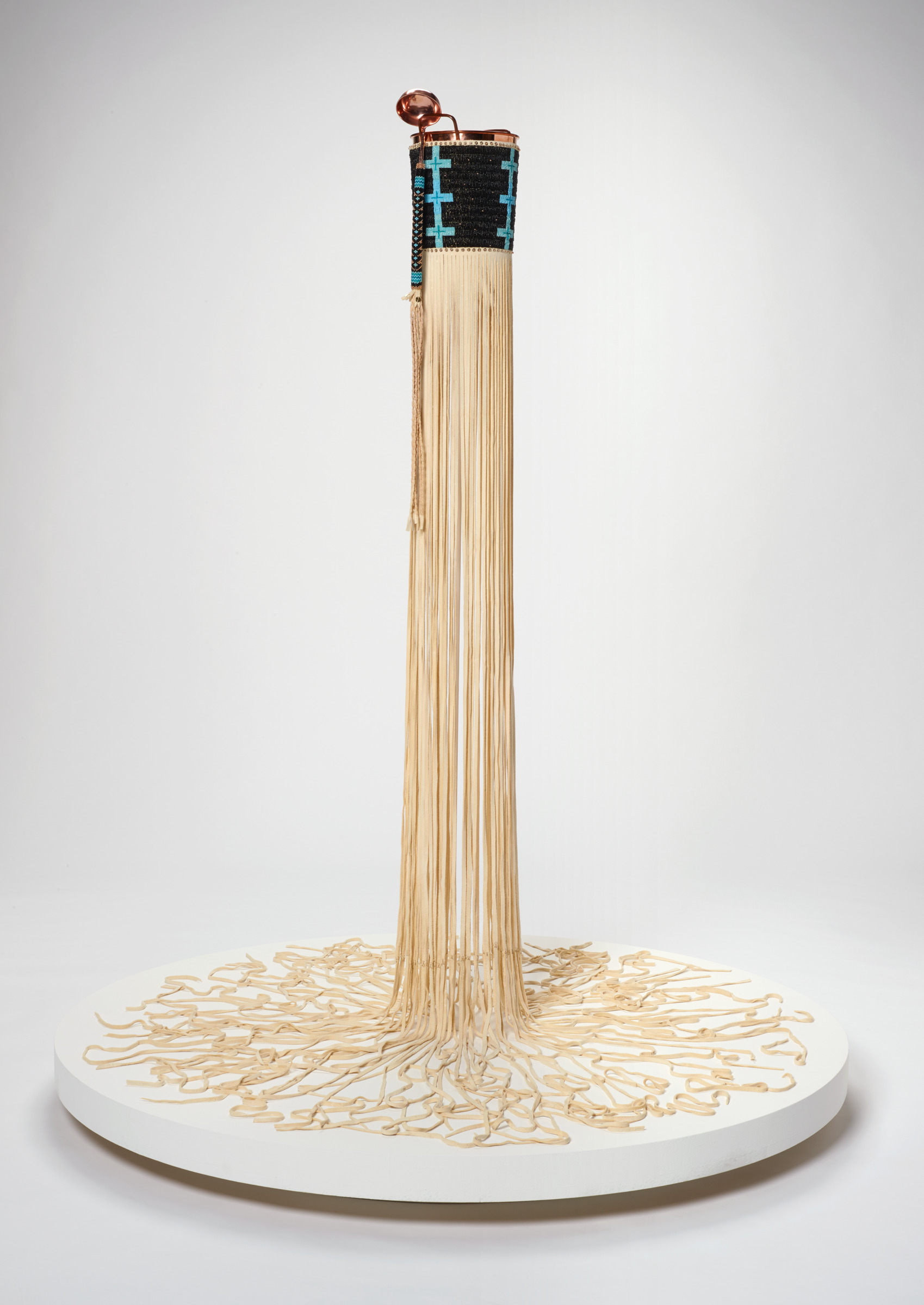 at the upper center of the image a copper bucket and ladle covered in black and blue beadwork are visible, from the bases of the bucket is a cascade of cream colored buckskin fringe which runs vertically onto a white circular platform base. The fringe radiates outwards in squiggling directions from the center
